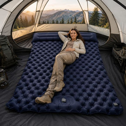 Matelas gonflable 2 personnes avec oreillers intégrés, installé dans une grande tente de camping, avec une femme souriante en tenue de randonnée profitant d’un couchage confortable en pleine nature montagneuse.