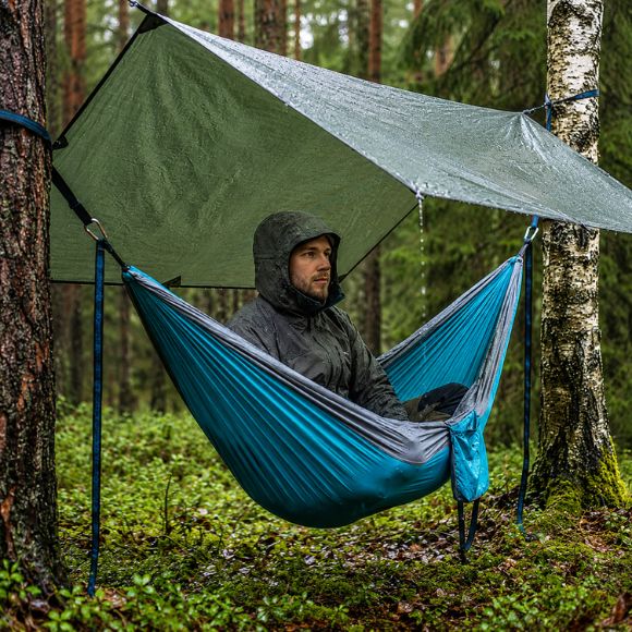 Hamac suspendue de camping avec bâche de protection installé entre deux arbres, utilisé par un campeur assis à l'abri de la pluie dans une forêt.